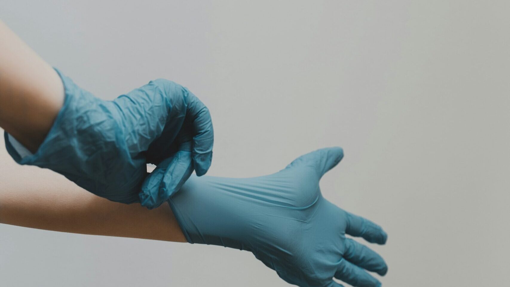 Eine Person zieht Einmalhandschuhe an. Zu sehen sind nur die Hände, welche von der Seite vor einem grauen Hintergrund fotografiert worden sind.